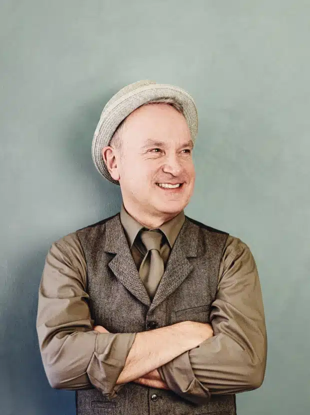 Ein lächelnder Mann mit hellgrauem Hut, braunem Oberhemd und dazu passender brauner Weste und Krawatte steht mit verschränkten Armen vor einem gedämpften grünen Hintergrund.