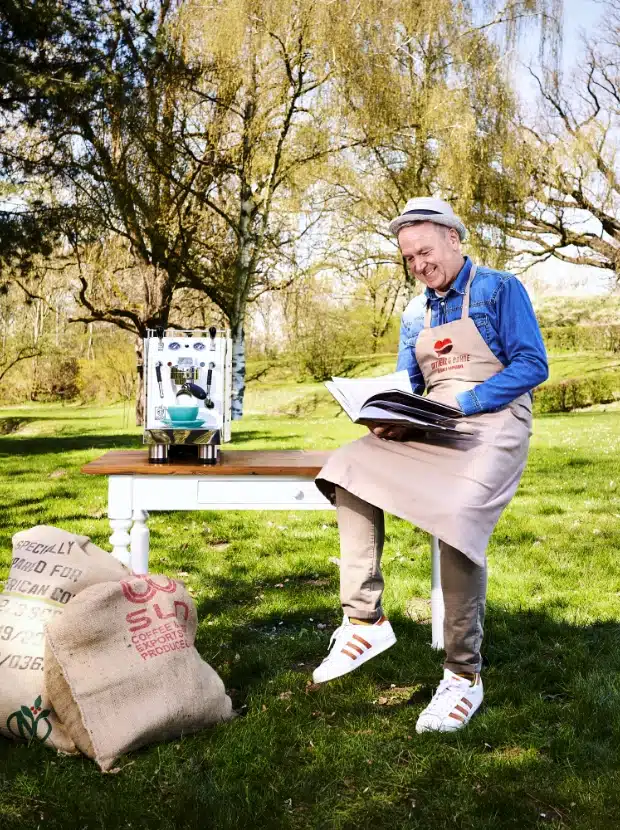 Ein Mann mit Schürze sitzt auf einem Holztisch in einer grasbewachsenen Umgebung mit Bäumen. Er lächelt und hält ein offenes Buch in der Hand. Neben ihm stehen eine Kaffeemaschine und mehrere Leinensäcke mit Kaffee-bezogenen Texten. Er wirkt zufrieden und entspannt.