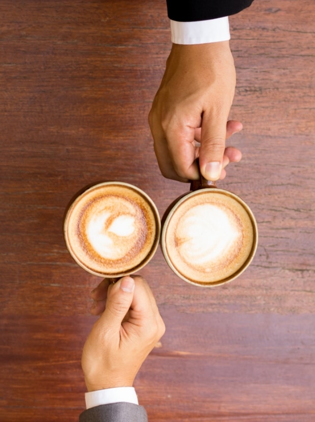 Zwei Hände, eine im schwarzen Anzug, die andere im grauen, halten Tassen Kaffee mit Latte Art auf einer Holzfläche. Die Tassen stehen dicht beieinander, von oben betrachtet.