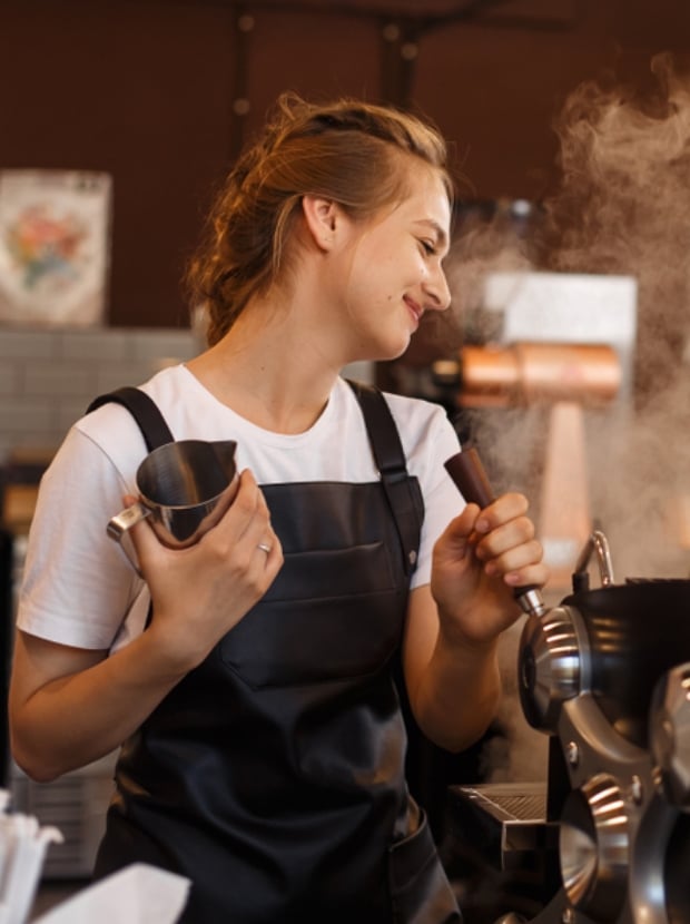Eine Barista mit zurückgebundenem Haar lächelt, während sie in einem Café Milch aufschäumt. Sie trägt ein weißes T-Shirt unter einer schwarzen Schürze und hält in einer Hand einen Krug, mit der anderen bedient sie eine Dampfdüse. Dampf steigt aus der Maschine auf.