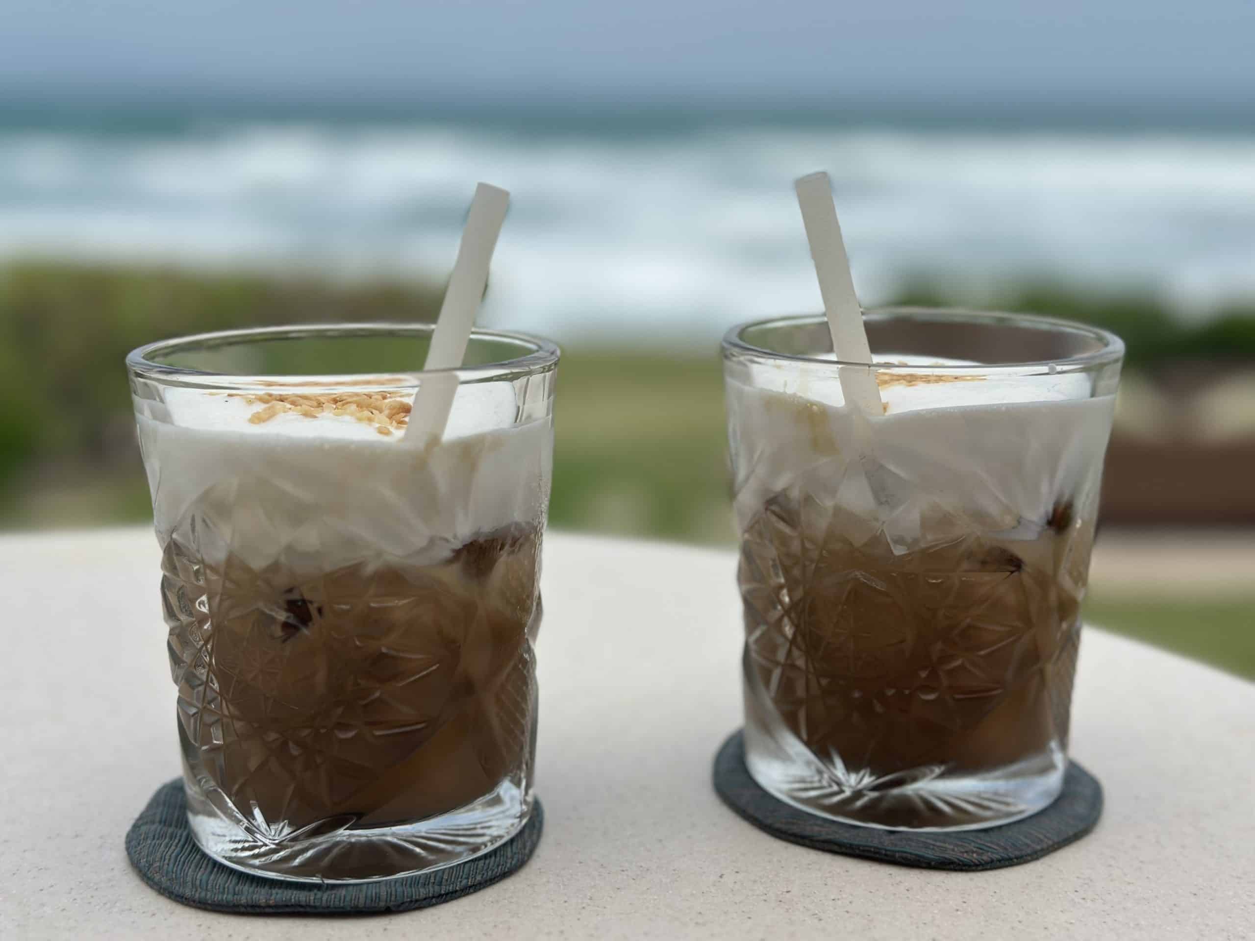 Zwei schaumige Eiskaffeegetränke in strukturierten Glasbechern mit weißen Strohhalmen stehen auf einem Tisch mit grauen Untersetzern. Im Hintergrund ist ein verschwommener Blick auf rollende Meereswellen unter einem bewölkten Himmel zu sehen, der zu einer entspannten Küstenatmosphäre beiträgt.