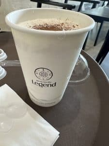 Ein weißer Pappbecher mit einem Trung Nguyen Legend-Logo steht auf einem dunkelbraunen Tisch. Der Becher ist mit einem schaumigen Getränk gefüllt, das mit Kakaopulver garniert ist. Daneben liegen einige Plastikdeckel, Besteck und eine weiße Serviette. Im Hintergrund sind Stühle und ein Tisch zu sehen.