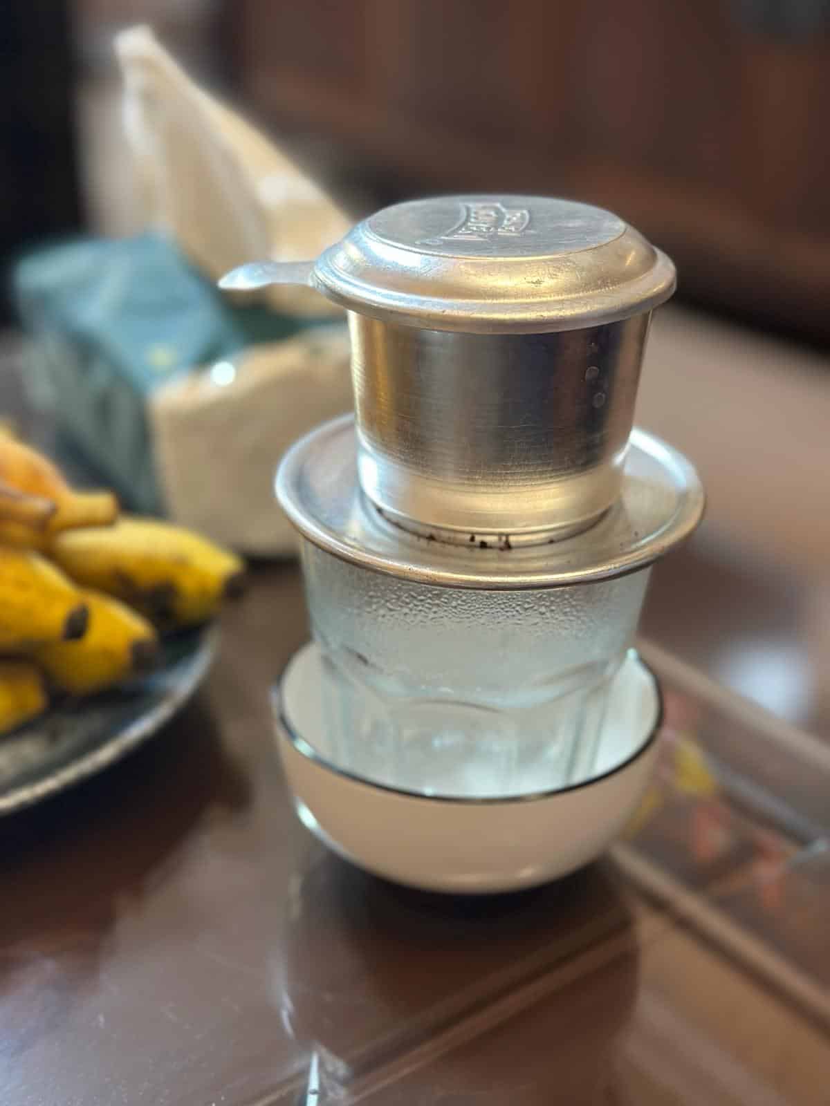Ein traditioneller vietnamesischer Kaffeefilter liegt auf einem Glas in einer weißen Untertasse. Im Hintergrund sind ein Bündel Bananen und eine Packung Taschentücher leicht verschwommen. Das Ganze steht auf einem glänzenden Holztisch.